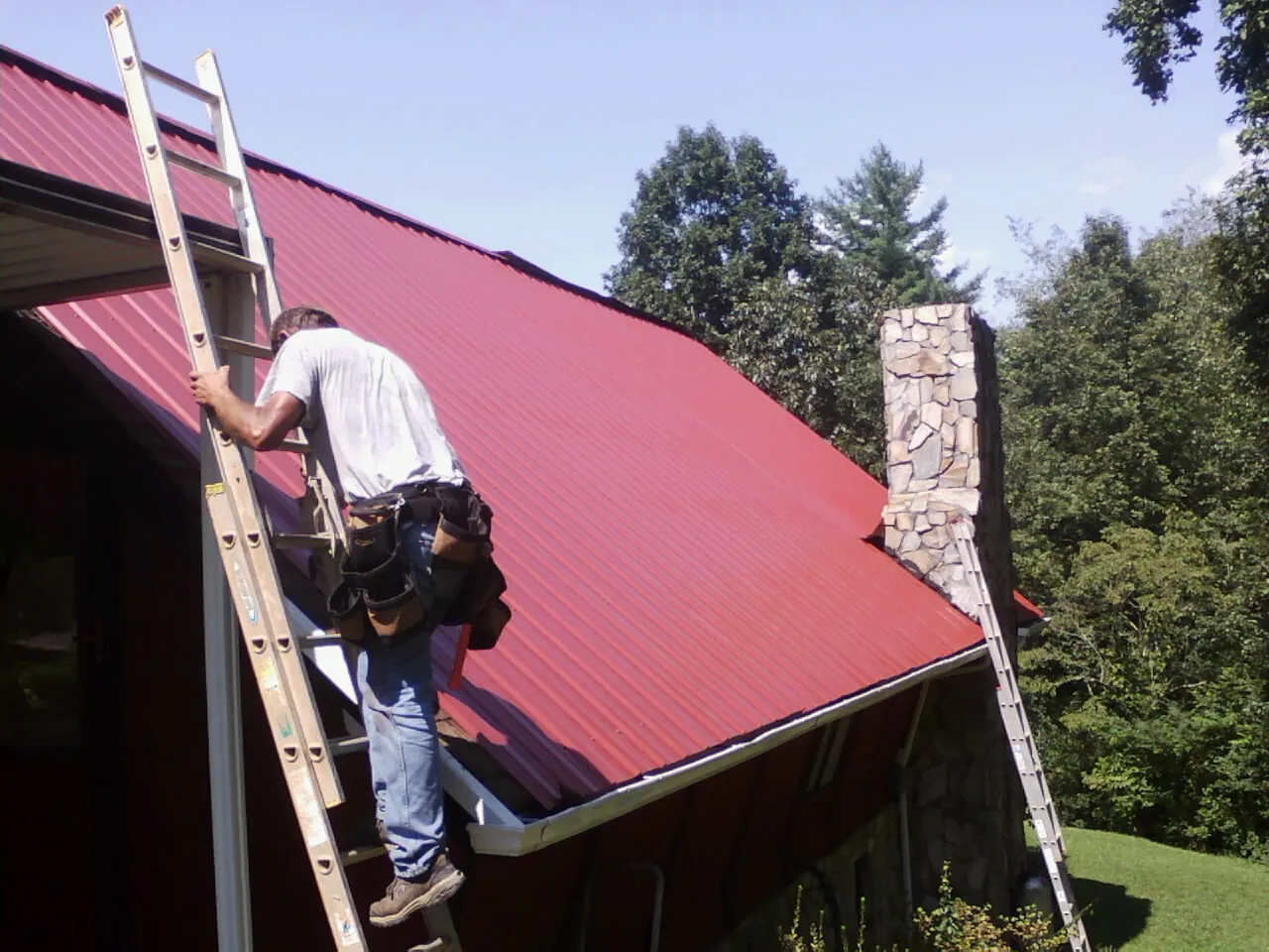Roofer on a ladder beside a red metal roof and stone chimney — Soffit & Fascia Repair by Roofing Brothers in Kuna